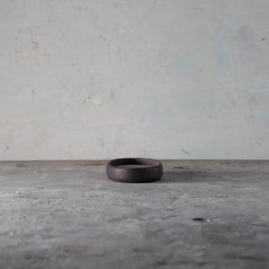 Puede incluir: Un pequeño plato circular de cerámica marrón oscuro está centrado en una superficie de hormigón gris. El plato tiene un perfil bajo con un borde ligeramente elevado. El fondo es una pared moteada de color claro.