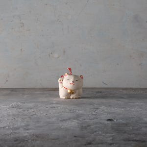 May include: A small, off-white ceramic Maneki Neko figurine with a raised paw, a red ribbon, and a gold bell. The cat has closed eyes and a smiling expression. The background is a mottled gray.
