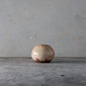 Puede incluir: Un jarrón esférico de cerámica con un esmalte beige claro y una base marrón oscuro. El jarrón tiene forma redondeada y una pequeña abertura en la parte superior. El fondo es una pared gris claro texturizada.