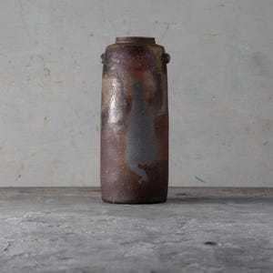 Puede incluir: Un jarrón de cerámica alto y cilíndrico con cuello estrecho y asas pequeñas. El jarrón tiene una paleta de colores moteados, terrosos, marrón, óxido y gris, con un brillo metálico. El jarrón se exhibe sobre una superficie de hormigón.