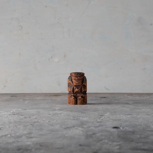 May include: A small, carved wooden figure of a seated person. The figure is brown with dark brown carved details, and it is positioned on a gray surface against a light background. The carving has a geometric design.