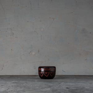 Puede incluir: Un pequeño recipiente redondo de color marrón oscuro con tapa. El recipiente tiene un patrón geométrico en forma de chevrón en color marrón rojizo. Está sobre una superficie gris sobre un fondo gris.
