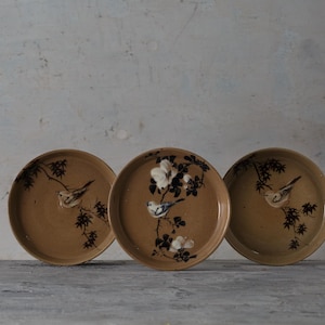 May include: Three round ceramic plates with a tan finish, each featuring a hand-painted design of a small bird perched on a dark brown branch. The center plate includes white floral accents, while the two outer plates show birds on branches.