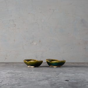 May include: Two small, handmade ceramic bowls with an organic shape. The bowls have a glossy, dark green and teal glaze, and sit on small, white feet. The background is a textured, light grey surface.