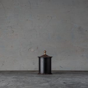 Può includere: Un contenitore cilindrico nero con coperchio e pomello dorato. Il contenitore è posto su uno sfondo grigio e su una superficie grigia. Il contenitore sembra essere in metallo.