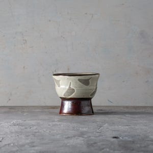 Pode incluir: Uma caneca de cerâmica artesanal com um design único. A caneca apresenta uma seção superior creme adornada com padrões cinza em forma de folha. A base é marrom escuro rico, e a borda também é marrom. A caneca está em uma superfície cinza.