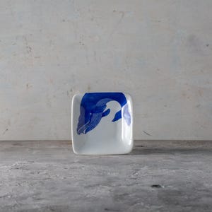May include: Square white ceramic dish with a blue whale design. The whale is depicted in a stylized, abstract form, with a curved body and detailed lines. The dish has rounded corners and a glossy finish.