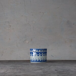 May include: A small, cylindrical ceramic container with a lid. The container is white with a blue floral and geometric pattern around the body. The lid has a similar design. The container is sitting on a dark surface.