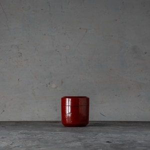 May include: A small, cylindrical, red container with a glossy finish. The container is set against a neutral grey background, with a dark grey surface below. The container appears to be made of wood or a similar material.