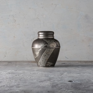 May include: A dark gray decorative jar with a lid, featuring etched silver-colored designs of trees and abstract shapes. The jar is cylindrical with a rounded top and sits on a gray surface.