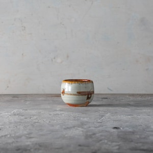 May include: A handcrafted ceramic cup with a rounded form. The cup has a white base with brown and orange streaks and a dark orange rim. The cup is set against a neutral backdrop.