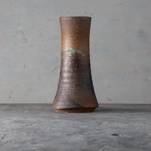 May include: A tall, handcrafted ceramic vase with a flared base and a cylindrical body. The vase has a textured, earthy brown finish with lighter brown and green accents. It stands on a gray surface against a neutral background.
