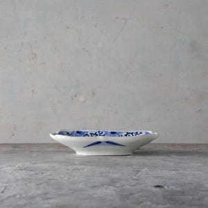 May include: A small, white ceramic dish with a blue floral design. The dish has a shallow, oval shape with slightly raised edges. The blue pattern is symmetrical and covers the rim and interior of the dish. The dish is set against a neutral background.