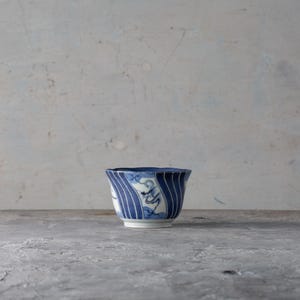 Puede incluir: Una taza de té de cerámica azul y blanca con borde acampanado. La taza presenta rayas azules verticales y un diseño de olas estilizadas. La taza está sobre un fondo neutro.