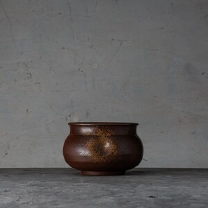 Puede incluir: Una olla de cerámica marrón con un cuerpo redondeado y un borde ligeramente ensanchado. La olla es de color marrón oscuro con motas marrones claras. Se asienta sobre una superficie gris contra un fondo gris.