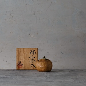 May include: A square wooden box with black Japanese calligraphy and a red stamp on the front, positioned next to a small, round, textured brown ceramic object resembling a piece of fruit with a green stem on top.