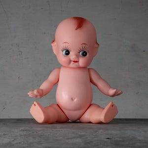 May include: A vintage-style baby doll with a pink body, sitting with its legs spread and arms outstretched. The doll has large eyes, long eyelashes, and a small red mouth. A small tuft of red hair sits atop its head.