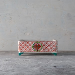 Op de afbeelding: Rechthoekige keramische plantenbak met een rood-wit ruitpatroon. De plantenbak heeft een centraal ontwerp met een ruitvorm en groene accenten. Hij staat op vier turquoise voetjes.