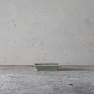 May include: A small, rectangular, pale green ceramic dish. The dish has a slightly flared shape and is centered on a grey surface. The background is a textured, off-white wall.