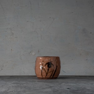 May include: A handmade, small, round, ceramic pot with a textured, brown surface. The pot features dark brown floral designs. The pot is sitting on a gray surface against a gray background.