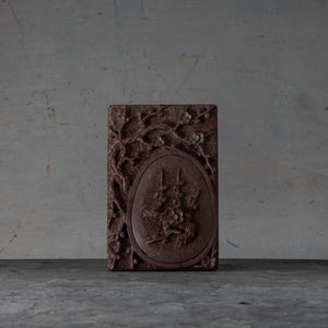 Pode incluir: Objeto decorativo retangular marrom-avermelhado com entalhes intrincados. O design inclui padrões florais e de galhos de árvores em torno de uma imagem central oval. A imagem central apresenta um relevo detalhado de uma cena com figuras.
