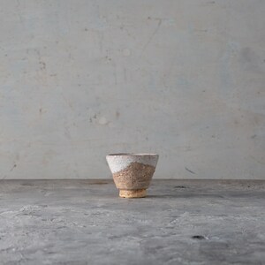 May include: A handmade ceramic cup with a unique, textured design. The cup features a white upper portion, a tan middle section, and a light brown base. The cup has a conical shape and is set against a neutral background.