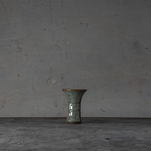 May include: A small, flared ceramic vase featuring a light sage green glaze with a prominent dark crackle pattern throughout the surface. The vase has a wide, open mouth that tapers down to a narrow base, showcasing a smooth, glossy finish.