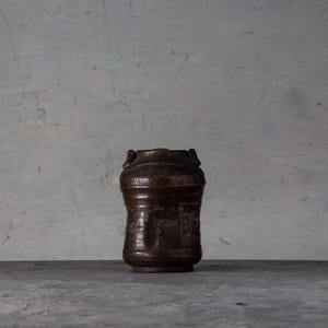 May include: A dark brown, handcrafted ceramic vase with a textured surface. The vase has a cylindrical shape with a slightly flared top and two small handles. It sits on a gray surface.