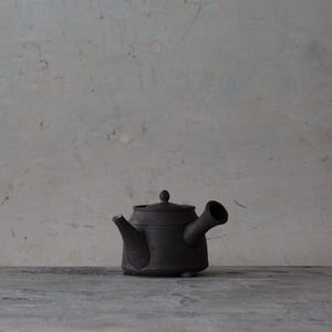 May include: A dark charcoal gray ceramic teapot with a side-mounted handle and a short spout. The teapot features a flat lid with a small knob handle and a low-profile, cylindrical body with a matte, textured finish.