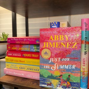 Könnte beinhalten: Eine Sammlung farbenfroher Bücher von Abby Jimenez auf einem Holzregal. Zu den Titeln gehören "Just for the Summer", "Yours Truly" und "Say You'll Remember Me". Die Buchcover weisen lebendige Designs und Text auf.