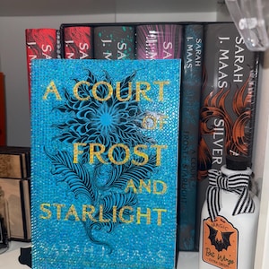 Könnte beinhalten: Ein türkisfarbenes Buch mit dem Titel "A Court of Frost and Starlight" mit goldfarbenen Buchstaben und einem floralen Design. Auf einem Regal sind weitere Bücher zu sehen, zusammen mit einer dekorativen Flasche mit einem Fledermausflügel-Etikett.