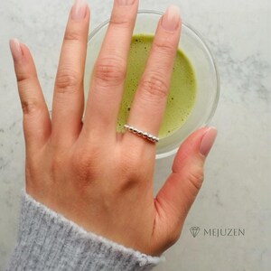 May include: A hand wearing a silver beaded ring is holding a glass bowl of green liquid, possibly matcha. The person is wearing a grey sweater. The brand name "MEJUZEN" is visible in the bottom right corner. The nails are painted with a light pink color.