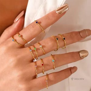 May include: A collection of gold-colored rings, each featuring a small, colorful gemstone. The gemstones are in various colors, including blue, green, red, and pink. The rings are displayed on a hand, showcasing their delicate design and vibrant accents.