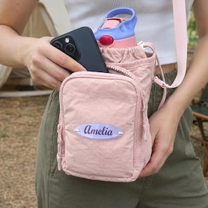 May include: A light pink crossbody water bottle holder bag with a front zipped pocket and a personalised oval name tag that reads Amelia in purple script. The bag features a drawstring closure and a matching pink adjustable shoulder strap.