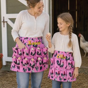 May include: Two pink egg-collecting aprons featuring a repeating rooster pattern and embroidered names. The aprons have multiple individual pockets designed to hold eggs securely. The fabric is a vibrant pink with black, white, and red rooster illustrations.