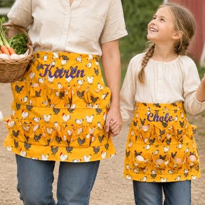 May include: Two mustard yellow aprons featuring a repeating chicken pattern and multiple pockets for egg collection. The larger apron is embroidered with the name Karen, and the smaller apron is embroidered with the name Chloe in purple script.