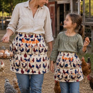 May include: Two egg-collecting aprons made of cream-colored fabric featuring a repeating pattern of colorful roosters. Each apron has multiple pockets holding brown eggs and is embroidered with the names Sarah or Lucy in orange thread.