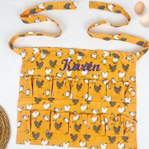 May include: A mustard yellow apron featuring a repeating pattern of white and black chickens. The apron includes two rows of multiple pockets and long fabric ties. The name Karen is embroidered in purple script across the center of the apron.