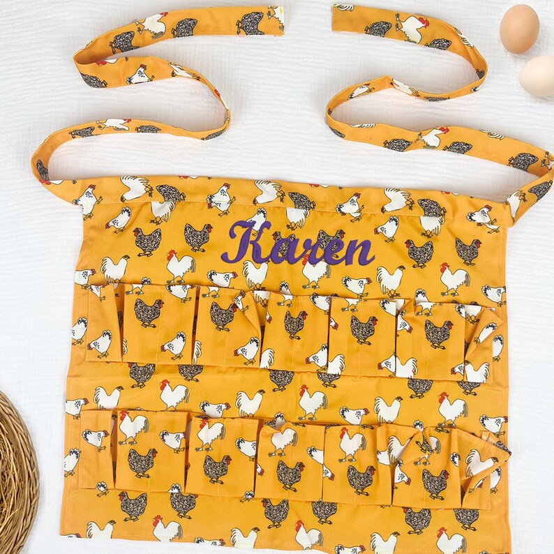 May include: A mustard yellow apron featuring a repeating pattern of white and black chickens. The apron includes two rows of multiple pockets and long fabric ties. The name Karen is embroidered in purple script across the center of the apron.