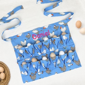 May include: A light blue egg collecting apron featuring a repeating pattern of white and brown chickens. The apron has multiple pockets, each holding a brown egg, and the name Daniel is embroidered in bright pink thread across the center.