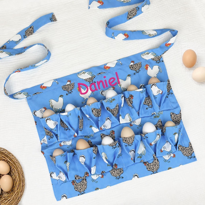 May include: A light blue egg collecting apron featuring a repeating pattern of white and brown chickens. The apron has multiple pockets, each holding a brown egg, and the name Daniel is embroidered in bright pink thread across the center.
