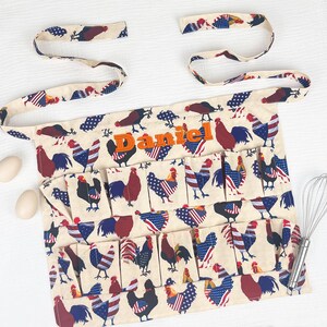 May include: A cream-colored apron featuring a repeating pattern of roosters with red, white, and blue American flag-inspired plumage. The apron includes multiple pockets and the name Daniel embroidered in orange thread across the upper chest area.