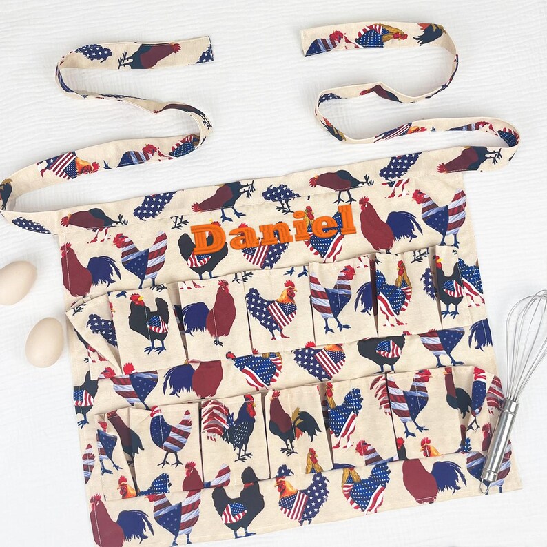 May include: A cream-colored apron featuring a repeating pattern of roosters with red, white, and blue American flag-inspired plumage. The apron includes multiple pockets and the name Daniel embroidered in orange thread across the upper chest area.