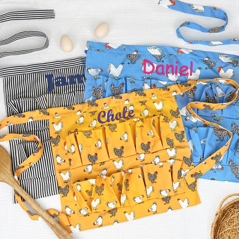 May include: Three waist aprons featuring chicken patterns in black and white stripes, blue, and yellow. The blue apron is embroidered with the name Daniel, the yellow apron with Chole, and the striped apron with Jamie. Each apron includes multiple pockets.