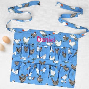 May include: A light blue apron featuring a repeating pattern of white and brown chickens. The apron has two rows of multiple pockets for holding eggs and is personalized with the name Daniel embroidered in bright pink script across the center.