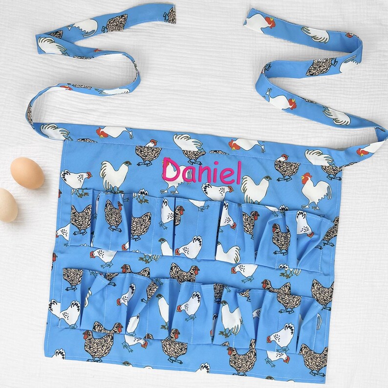 May include: A light blue apron featuring a repeating pattern of white and brown chickens. The apron has two rows of multiple pockets for holding eggs and is personalized with the name Daniel embroidered in bright pink script across the center.