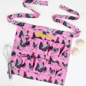 May include: A pink apron featuring a repeating pattern of black and white roosters with red accents. The apron includes multiple front pockets and the name Maria embroidered in yellow thread. A metal whisk and two eggs are placed nearby.
