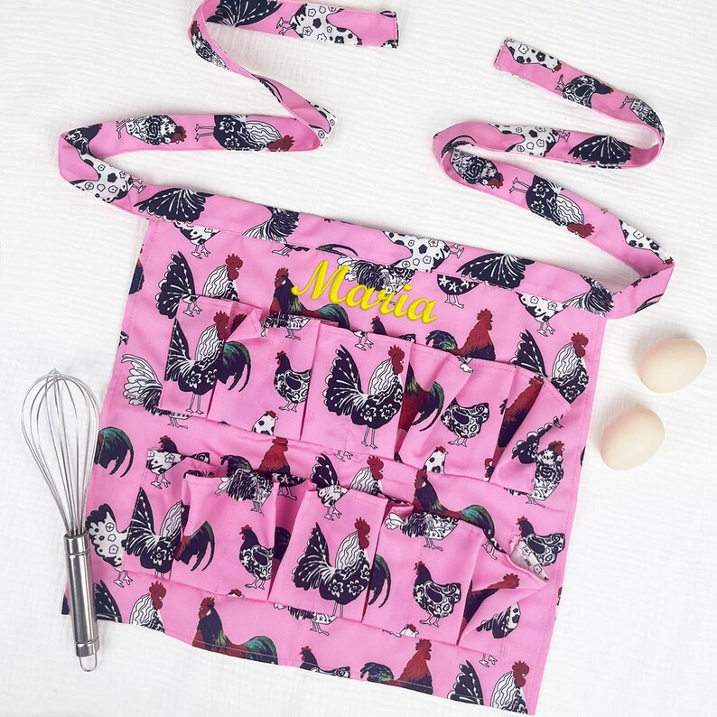 May include: A pink apron featuring a repeating pattern of black and white roosters with red accents. The apron includes multiple front pockets and the name Maria embroidered in yellow thread. A metal whisk and two eggs are placed nearby.