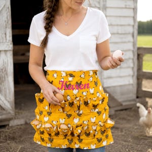 May include: A mustard yellow apron featuring a repeating pattern of white and black chickens, with multiple exterior pockets designed for holding eggs. The name Karen is embroidered in bright pink script across the center of the apron.