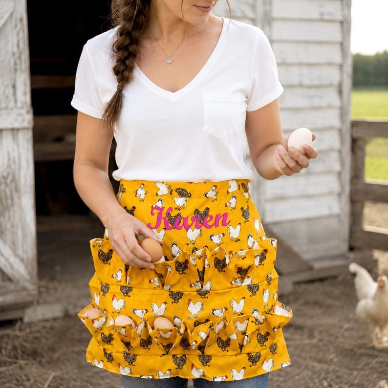 May include: A mustard yellow apron featuring a repeating pattern of white and black chickens, with multiple exterior pockets designed for holding eggs. The name Karen is embroidered in bright pink script across the center of the apron.